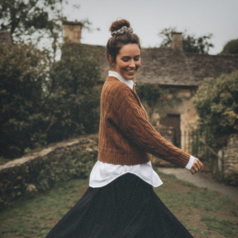 Woman in a brown sweater and black skirt standing outdoors with a stone building in the background, fawn suede scrunchie
